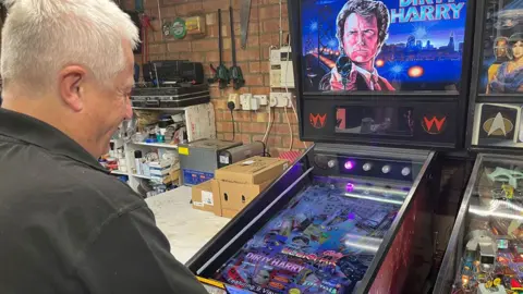 Jozef Hall/BBC A side view of Mark Squires with short white hair, wearing a grey polo shirt, in front of a Dirty Harry pinball machine