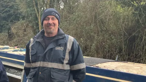 A man in a navy blue hat and blue windbreaker with hi-vis strips stands in front of a blue narrowboat.