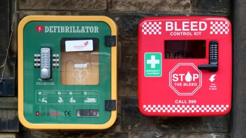 The defibrillator is in a square green and yellow cabinet, and the bleed kit in a square red cabinet. Both the boxes have been installed on a wall.