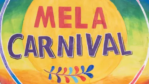 getty A painted canvas, it has bright colours. The backdrop is a gradient of blue, green into yellow and orange. In large letters it says 'Mela Carnival' in red and purple.