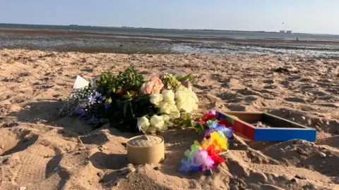 Henry Godfrey-Evans/BBC Flowers, a trinket box and paper wreaths left on the sand. The sea is in the distance. 