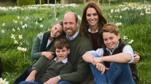 The Prince and Princess of Wales with their three children sitting on the grass amongst some daffodils. Prince William is at the centre alongside Catherine. He is balding with short brwn hair and a cropped beard, and is wearing a green jumper with a light blue shirt collar visible. Prince Louis, who has a gapped toothed smile, is wearing similar clothes and is sitting in his lap. Leaning on his right shoulder is Princess Charlotte who is wearing a green jumper and dark tartan-style scarf. She has long brown hair and is siling at the camera. On his left shoulder is his wife, Princess Catherine, who is smiling. She has long brown hair and is wearing a deep red jumper. Her arm is round Prince George who is in blue jeans, a brown gilet and white shirt with his sleeves rolled up.