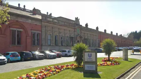 Google A red and yellow brick building with cars parked in the front. A grass area is planted with flowers and two small bushes. A sign reads Unilever Port Sunlight, Lever House.