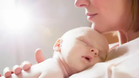 A baby sleeping on his or her mother's chest. The mum's chin, nose and lips can be seen with her chin gently resting on the baby's head. She wears a light top and has a blonde bob. 