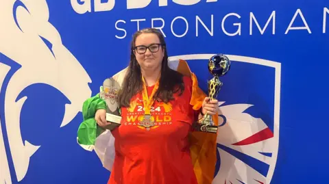 Erin Harris has long brown hair and glasses. She has trophies in both hands and is wearing a red t-shirt and an Ireland flag over her shoulders. There is a blue background behind her.