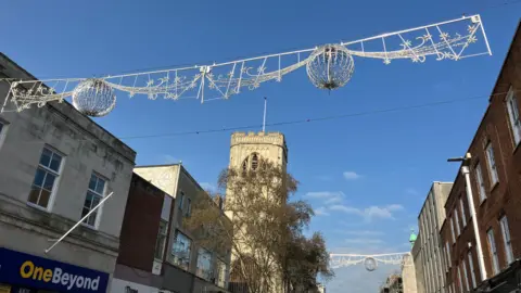 Silver Christmas decorations - wire banners with ball-like structures and swirls - hang above a city centre street on a sunny day in the spring. There is a church tower on the corner, and shops line the street.
