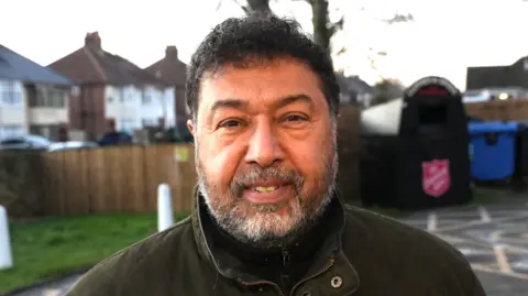 John Fairhall/BBC A man with short black hair and grey facial hair. He is smiling into the camera.