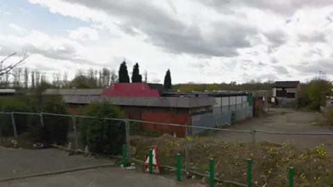 Google A large single storey building, surrounded by temporary perimeter fencing