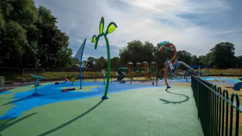 A water play area including structures that might be found on a children's playground. Trees are on the far left and railings are on the far right.