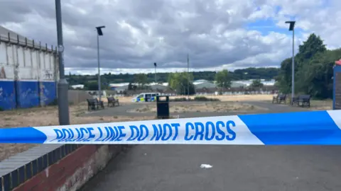 Blue-and-white police tape with the words "Police line do not cross". Beyond the tape, slightly out of focus, there is a litter bin and a circular path around a patch of grass. There are benches on either side of the path. On the left, there is a blue and white building with peeling paint. There are streetlights on poles. In the distance there is a police van.