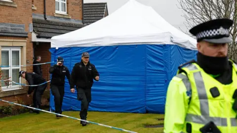 Getty Images Police officers outside a red-brick house. there is a large police tent on the lawn, with officers in all-black uniform walking out of the house across the lawn towards an officer in reflective uniform. 