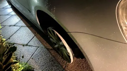 A silver car missing a tyre on its front wheel. The alloy wheel has been scuffed and worn down around the edges where it has been driven on. It is parked on concrete slabs, which look to be part of a drive