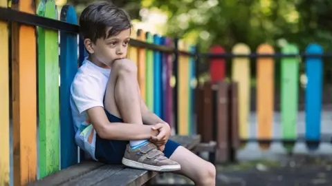 A young boy wearing a white T-shirt and navy shorts stares into the distance as he sits alone on a bench in a playground. The fence around him is painted in lots of different colours.