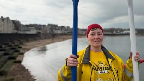 A women stands in front of the beach, you can see the coastline and buildings behind her. She is wearing a red hat and a yellow waterproof high vis jacket. she is also holding two oars the one in her right hand is blue the one in her left is white