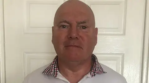 Philip Miller, who has a bald head and is wearing a white shirt with a dark red and black checked collar, is standing in front of a white door in a house. He looks impassive. 