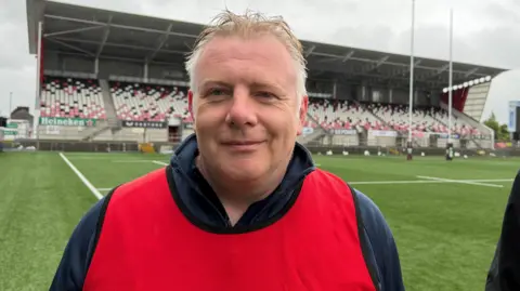 Šedý vlasy v červeném bryndáčku a námořnictvo polovinu zipu stojícího před ragbyovým sloupkem a posezením s hřištěm za ním.