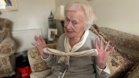 An older woman sitting on her couch holds up the bow in her shirt. She has white hair and blue eyes, and a crutch resting against her as she sits.