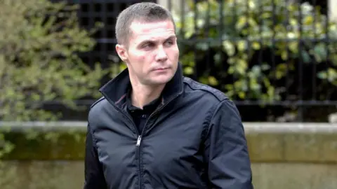 Spindrift A man with short cropped hair and a serious expression photographed outside a court building. He is looking off to his left.