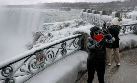 Joe Raedle/Getty Gambar Seorang wanita dengan syal merah di atas wajahnya dan mantel hitam dengan kap mesin mengambil selfie dari platform yang menghadap ke Air Terjun Niagara. Air terjunnya beku. Ada tanda pada pagar di atas platform yang mengatakan 'bahaya'.