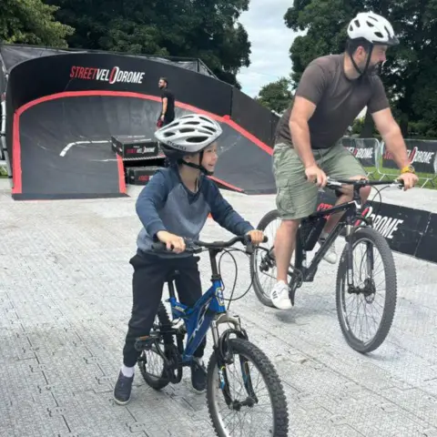 North Lincolnshire Council A man and a boy, both wearing casual clothes and helmets, ride bikes on a temporary cycle track. Behind them is a banked curve - a smaller version of those seen in velodromes - which is coloured black with red trim. It features the slogan "Street Velodrome". 