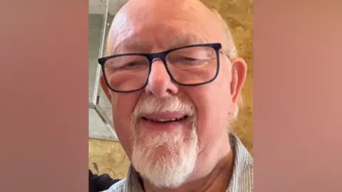 Graham Jackson A close up of a man with a white beard wearing black glasses, smiling down at a camera.