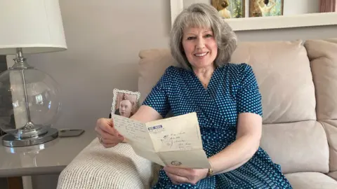 A woman with grey, white hair to her shoulders, sits in a light grey sofa with a table lamp to her right and a window behind her head. She wears a dark blue dress with light blue spots and holds a piece of folded paper in her hand with blue writing.