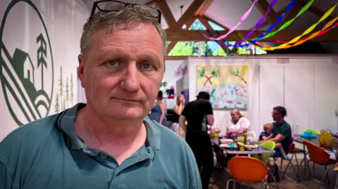 A man looks at the camera in a community hall with multi colours ribbons on the ceiling and people gathered around a table and chairs. he has short grey hair and is wearing a green t shirt