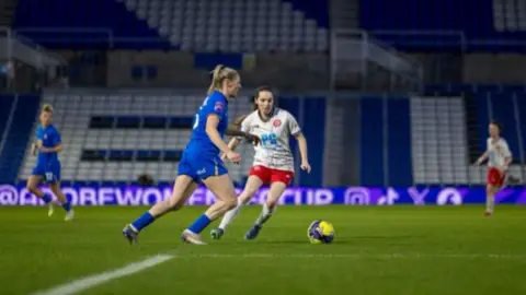 Players from Birmingham City and Chatham Town are on the pitch, with other team members in the background. There are football stands behind them.