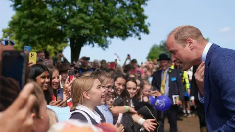 PA Media The Prince of Wales with schoolchildren as he attends the Royal Norfolk Show at the Norfolk Showground in Norwich. There is a large crowd of children many taking pictures on their phones. Prince William is wearing a blue suit and bending down slightly to talk to one of them. 