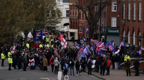 A large group of people carrying a mix of flags, including the Cross of St George and the Union flag, are outside Nuneaton Town Hall