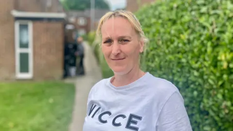 Nicola Rees/BBC A woman with blond hair wearing a grey top standing in front of a house with a hedge to her left