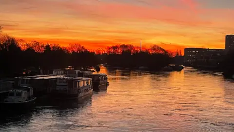 The sky is a beautiful yellow colour and turns dark orange as it hits the horizon. In the foreground, the river runs the breadth of the picture with several narrow boats moored up on the left-hand side. 