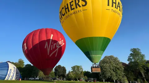 MJ Ballooning A Virgin-branded hot air balloon and a Thatchers -branded balloon hover over a playing field dotted with trees. A partially inflated blue and white balloon is visible to the left of the shot.