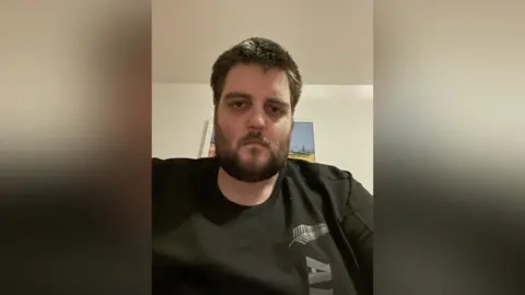 A man with dark hair and a dark beard looks into the camera. He is wearing a black t-shirt. 