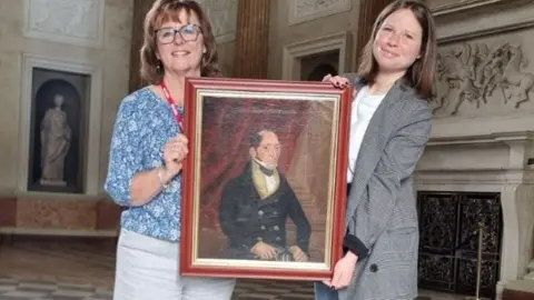 Two women hold a painting of a man in formal clothing. The women are stood in a grand room in a country house. 