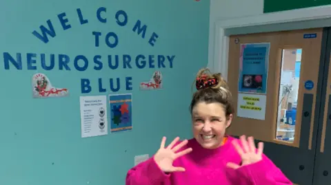 Brain Tumour Research Ms Wrathall smiles at the camera with her hands near her face in a wave. She wears a bright pink jumper. Behind her on a light blue wall, navy letters read: Welcome to Neurosurgery Blue.