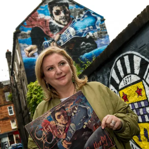 Fotografía digital pictórica Paisley Martha Rafferty se encuentra frente a un mural al costado del mismo mural que sostiene la portada del álbum de su padre.