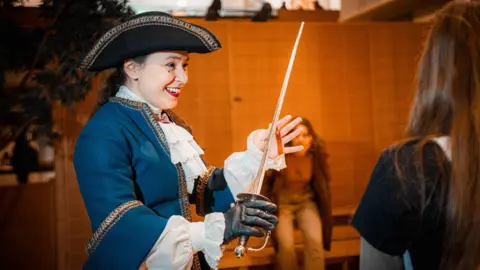 A woman in a historical French costume of a tricorn hat, blue jacket and white shirt smiles as she holds a sword.