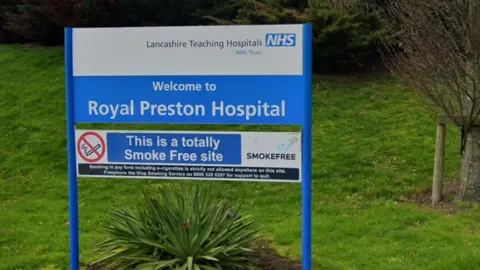 A blue and white sign reading 'Welcome to Royal Preston Hospital' in front of green bushes and grass