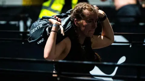 Ciarán Watts Suzanna performing walking lunges at a Hyrox competition. Her brown hair is braided and she is wearing a black sports top. She is carrying a black sandbag on her back and kneeling into lunges.