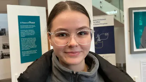 Isabella Zinkova is smiling and seen wearing clear-rimmed glasses and has her brown hair pulled back. She is wearing a grey fleece with a black coat. She is standing in the main atrium of the Ulster University campus in Derry. 