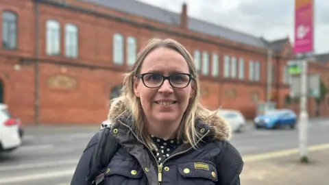 Jane Gill has blonde hair down to her shoulders and is wearing black glasses. She is also wearing a black puffer coat with a fur trim around the hood and has a black and white spotted top on.