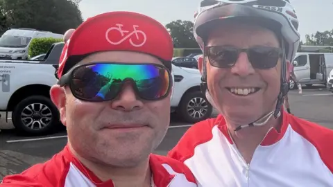 Most of the photo features two men, both wearing sunglasses and wearing white and red tops. Vehicles are in a car park behind them.