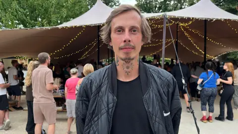 Steven Stone wearing a black T-shirt under a black jacket with a white Nike logo on the left chest. He has short brown hair and has tattoos on his neck. He is standing in front of the marquee in the Trinity Centre gardens.