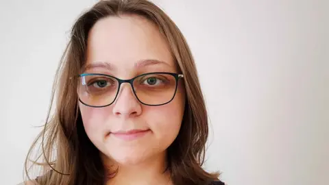 Ashley Walker A female wearing glasses, standing in front of a pale white background.