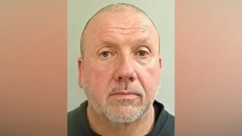 A mugshot of a middle aged man. He has a shaved head and a short grey beard. He is wearing a black T-shirt and black sweater jumper. 