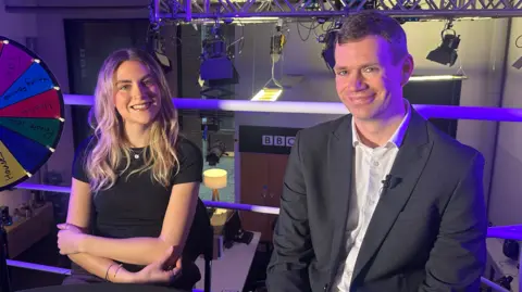 A female reporter and a male reporter look at the camera smiling