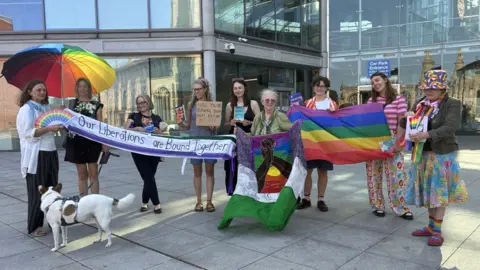 Zoe Dennis/BBC A line of nine people standing side by side outside a large glass building. A woman is holding a rainbow-coloured umbrella, another is holding a rainbow fan. Two are holding up a rainbow flag. A fabric banner reads Our Liberations Are Bound Together. Several are holding up books. 
