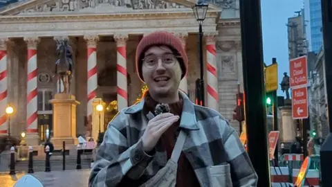 PA Media Tom wearing a red beanie hat, glasses and a checkered shirt while speaking into a microphone.