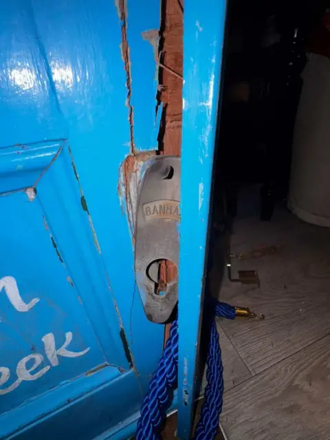 Close-up of a damaged bright blue wooden door with cracked paint and splintered wood around a metal lock plate. A piece of blue rope has been looped through the handle area.
